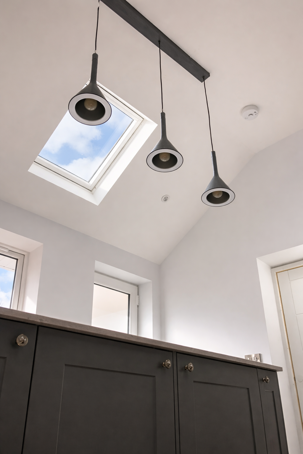 Interior ceiling with skylight showing blue sky and clouds, three hanging pendant lights, and two windows in the background.