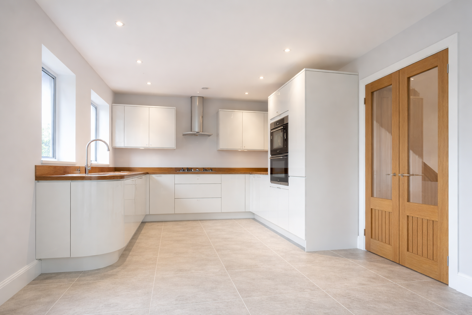 Modern kitchen with white cabinets, wooden countertops, stainless steel appliances, and beige tiled flooring. There are three windows and wooden double doors.