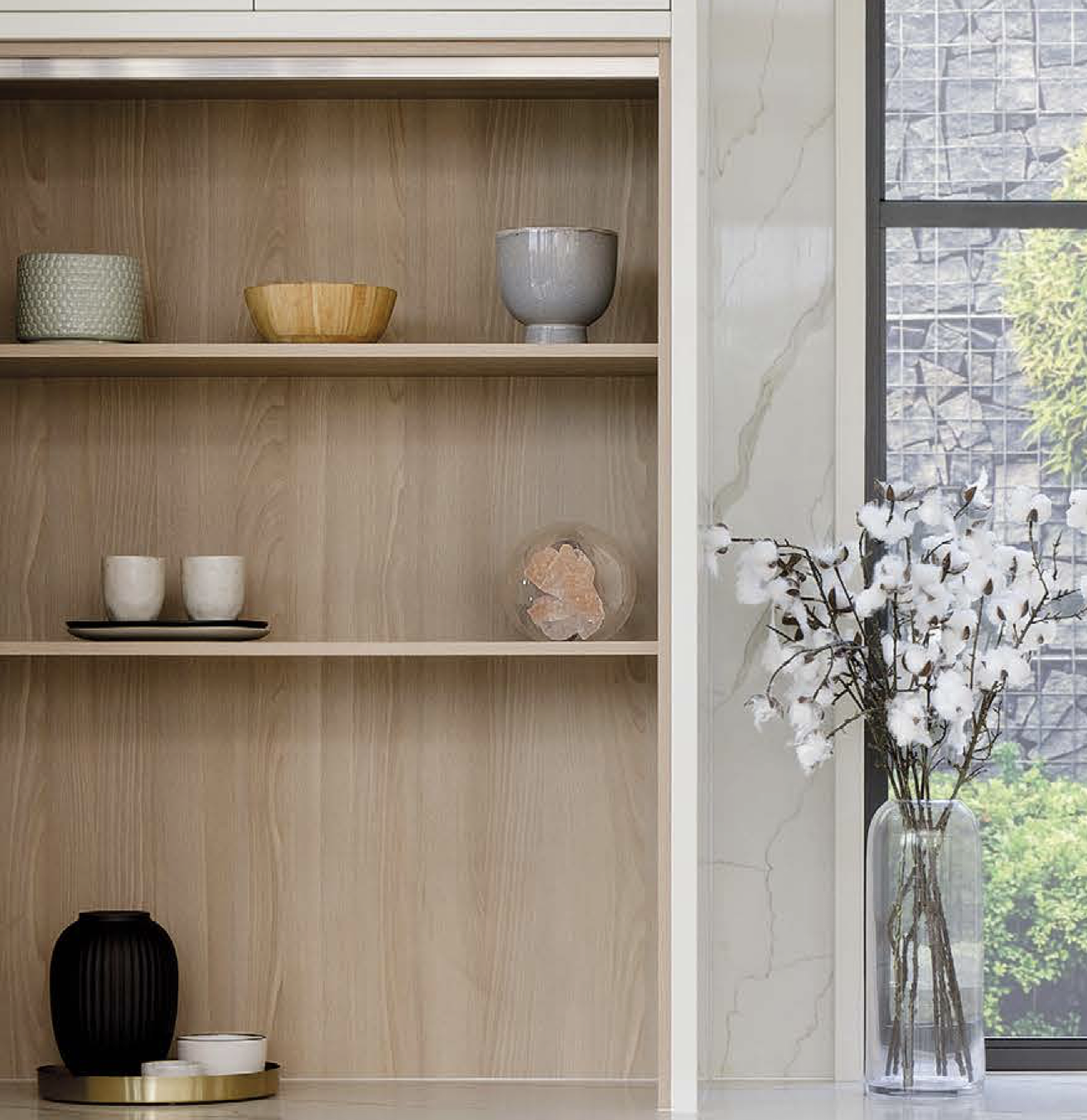 Wooden shelves with decorative ceramics next to a window with a vase of white cotton branch flowers.
