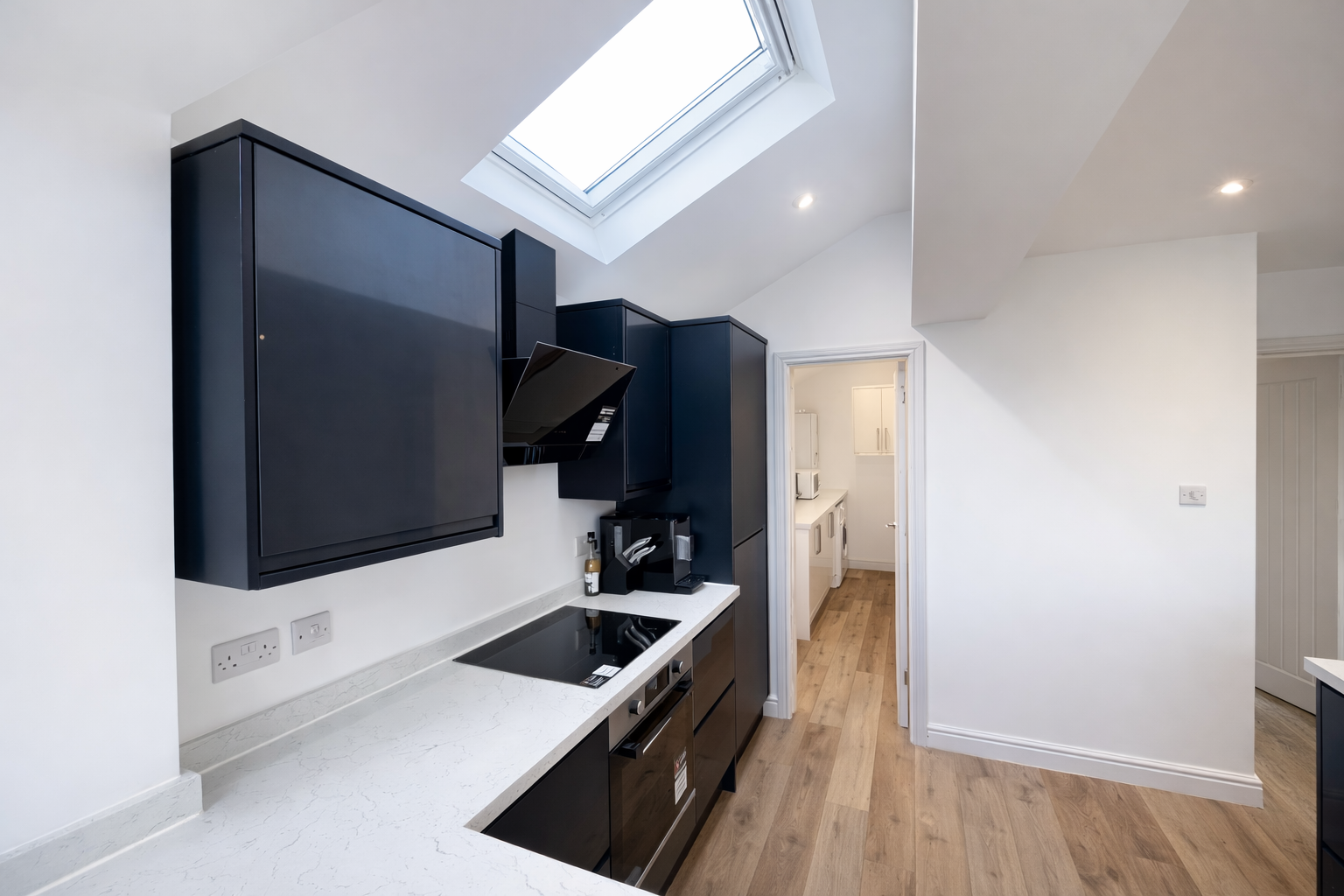 Modern kitchen with black cabinets, white marble countertop, built-in stovetop, coffee machine, and a skylight.