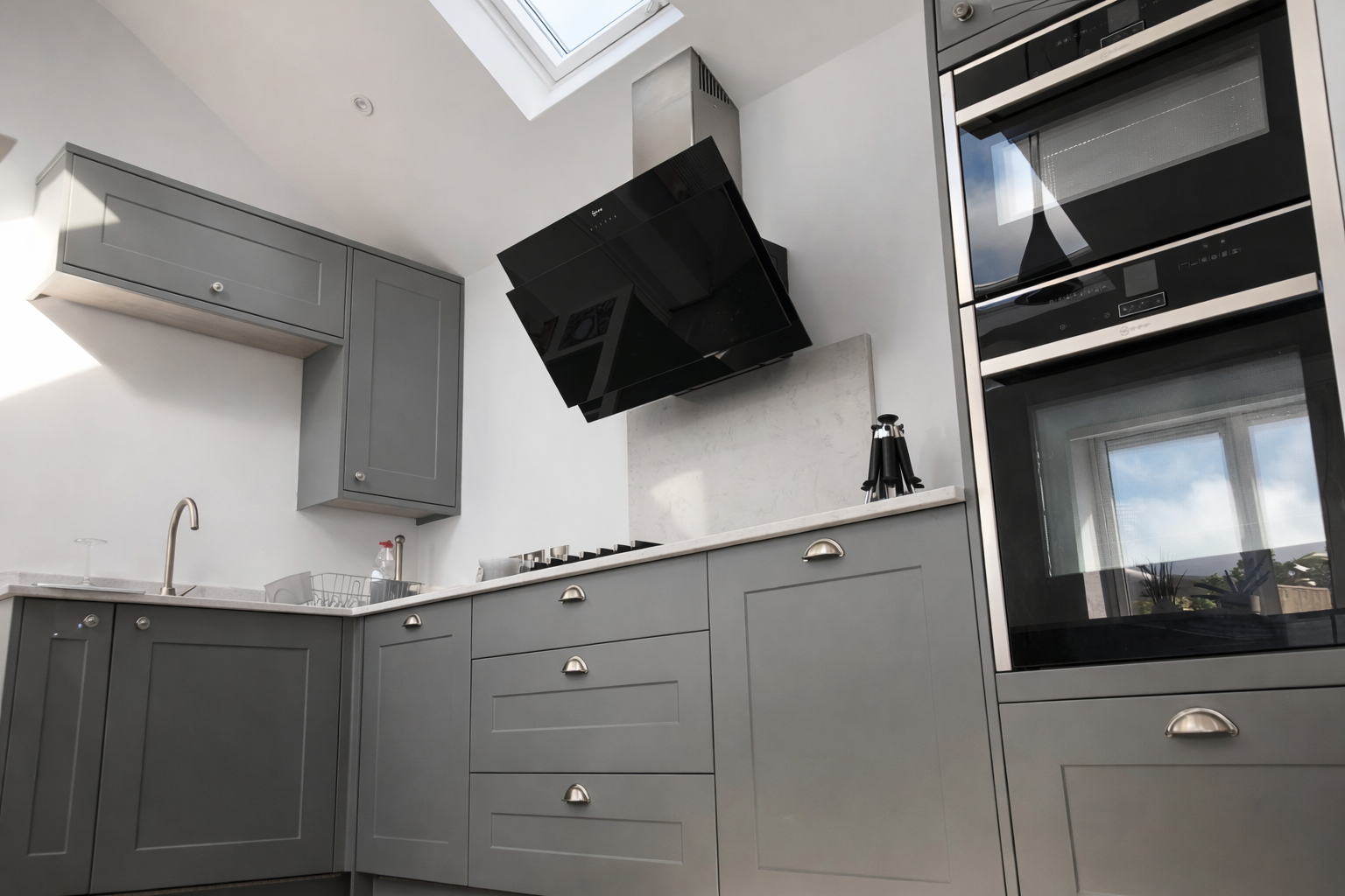 Modern kitchen with gray cabinets, black range hood, and built-in oven, sunlight coming through a window and skylight.