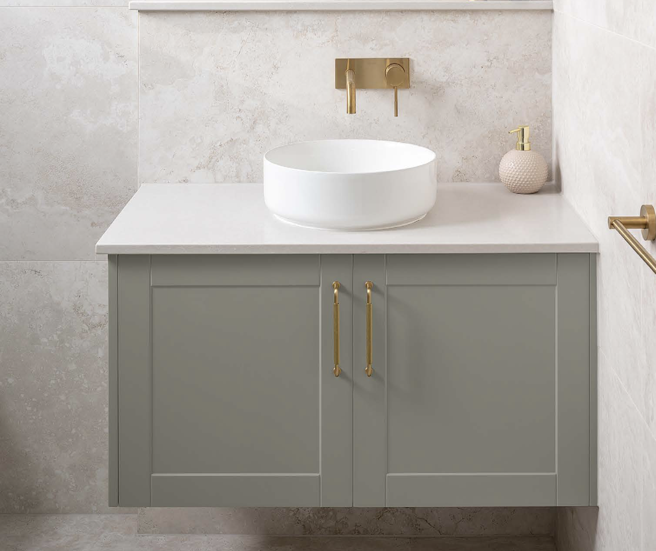 Bathroom vanity with a beige countertop, gray cabinet with gold handles, white vessel sink, wall-mounted gold faucet, pink textured soap dispenser, wall with beige textured tiles, and gold towel bar.