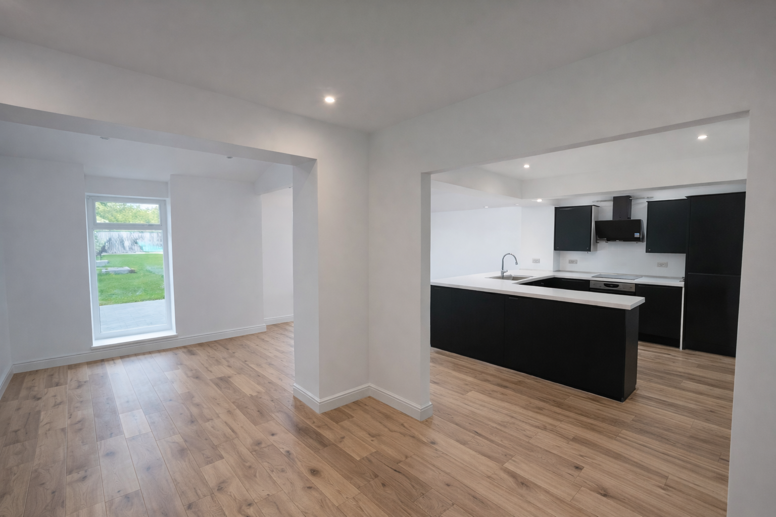 Empty modern kitchen with black cabinets and white countertops, open to a room with wooden floors and a large window.