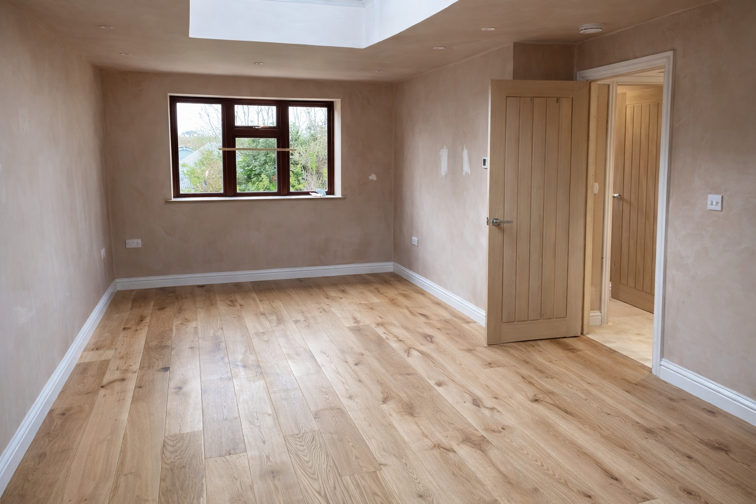 Empty room with wooden flooring, beige walls, a window with greenery outside, and a partially open wooden door leading to a hallway.