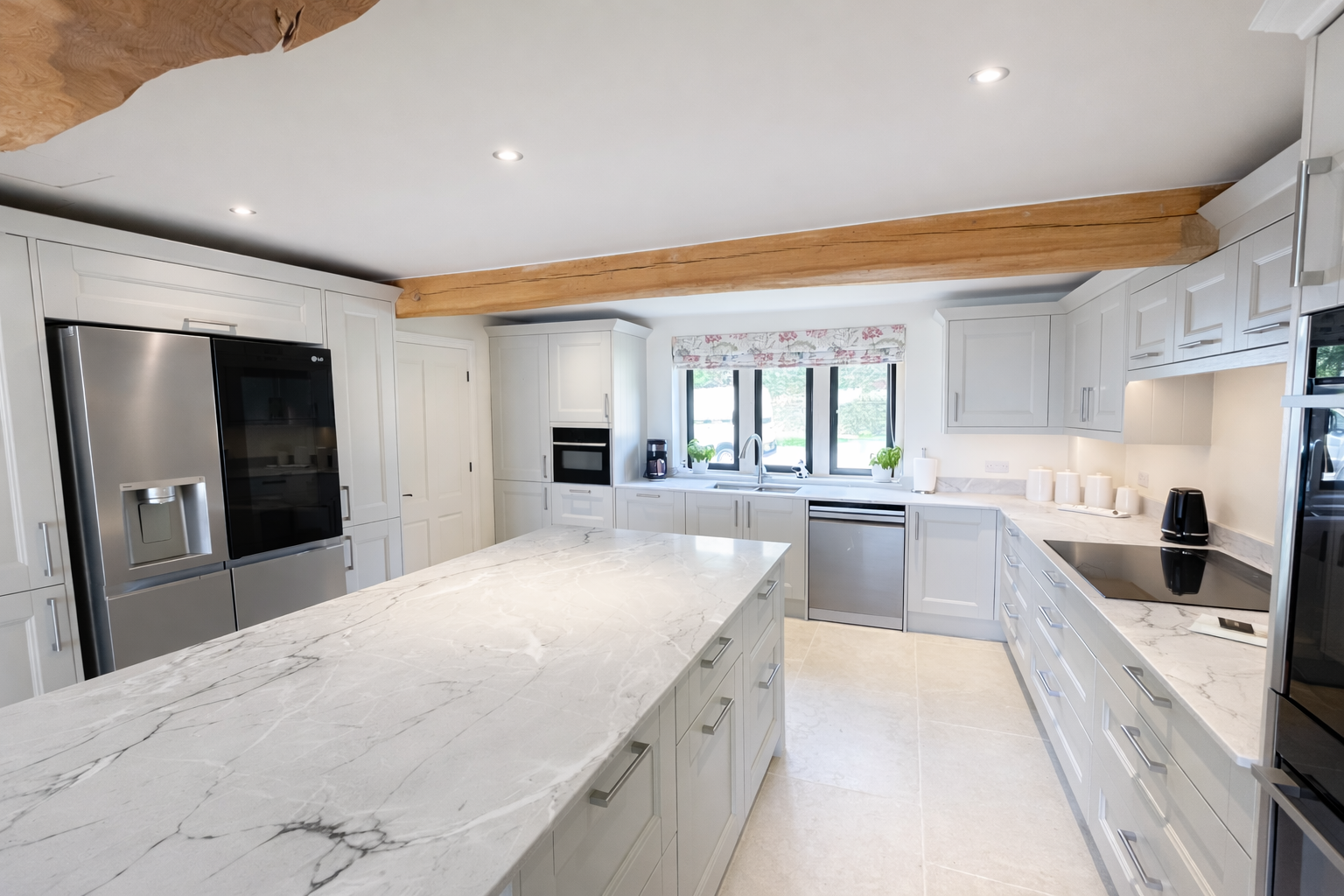 A spacious kitchen with white cabinets, marble countertops, a large kitchen island, stainless steel appliances, a window with floral valance, and wood ceiling beams.