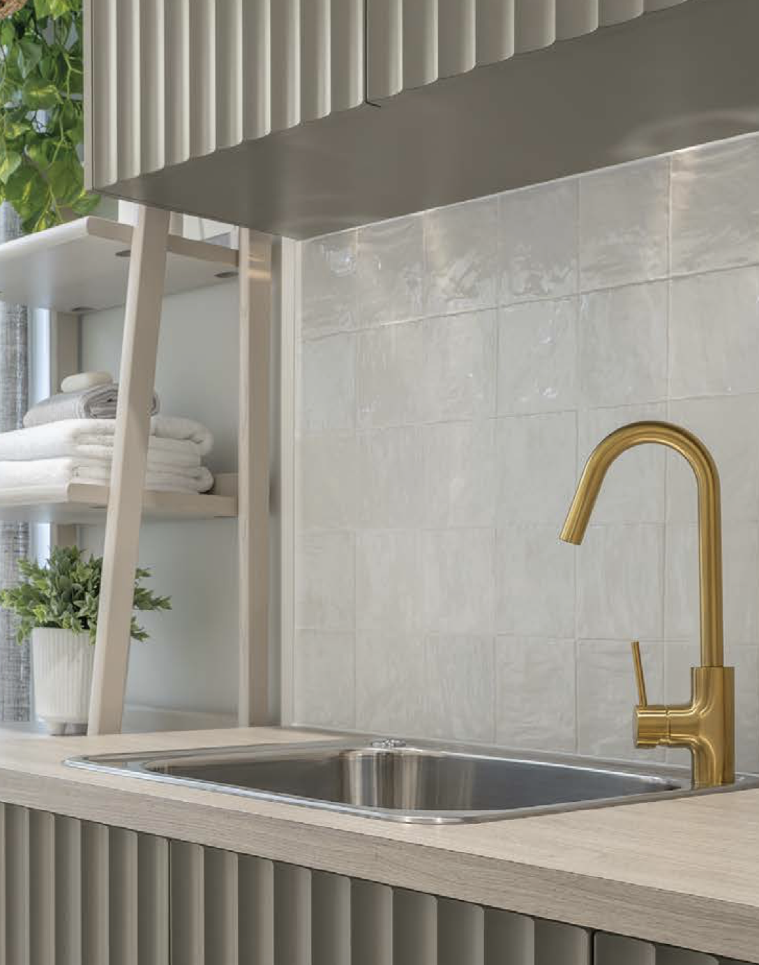Close-up of a kitchen sink with a gold faucet, beige textured backsplash, and a light wood countertop. In the background, there is a white shelf with folded towels and a potted green plant.