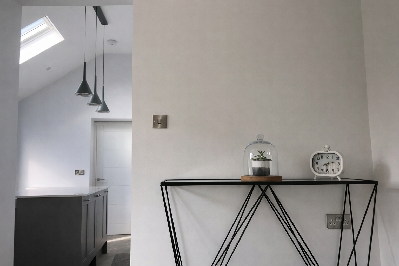 A minimalist interior with a black metal console table against a white wall, holding a small potted plant under a glass cloche and a white vintage-style alarm clock. In the background, a kitchen area with gray cabinets, a door, and three hanging black pendant lights is visible, along with a skylight window.