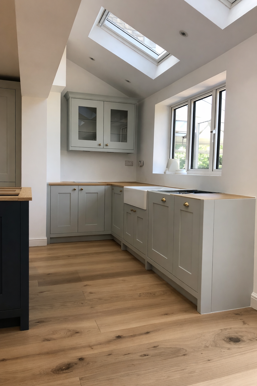 A kitchen with white cabinets, a farmhouse sink, wooden countertops, oak flooring, and large windows with a skylight overhead.