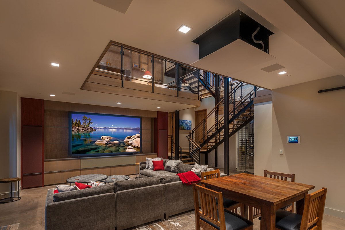 Living room with a large wall-mounted flat-screen TV showing a lake scene, a gray sofa with red and gray pillows, a wooden dining table with four chairs, a staircase with black metal railings, and a modern interior design.
