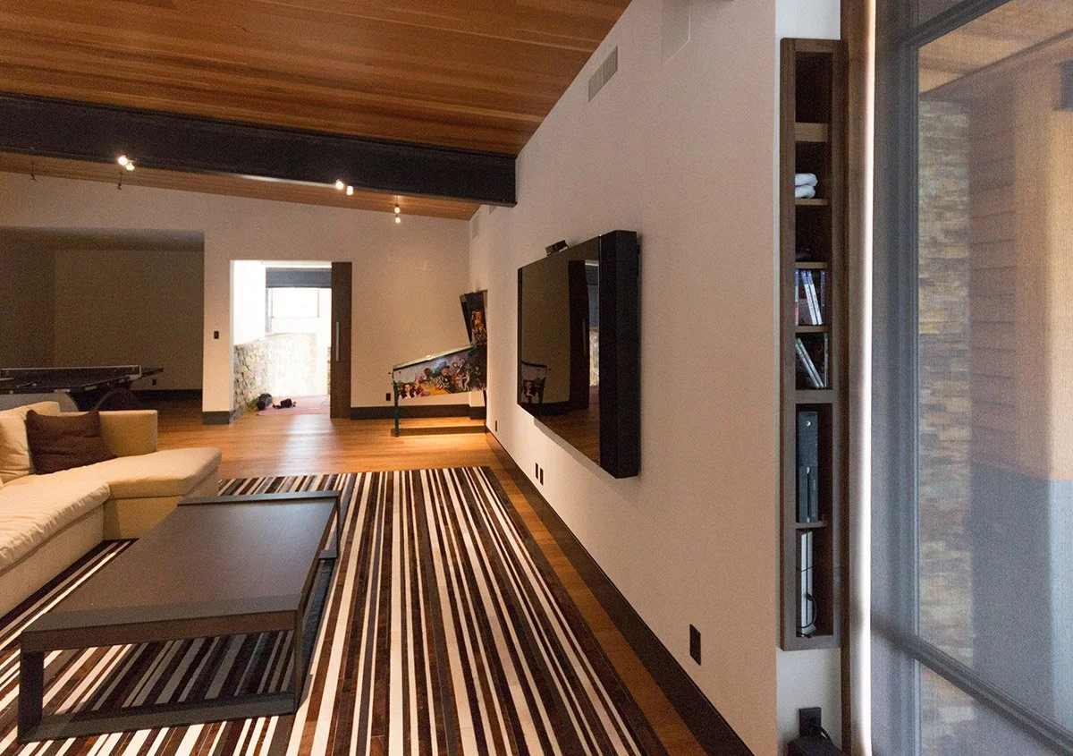 Modern living room with a white couch, striped rug, wall-mounted TV, built-in shelf with books, wood ceiling, and sliding glass doors.