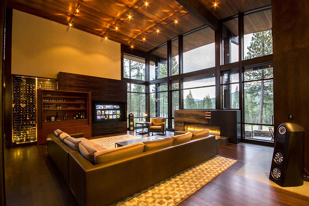 Living room with large floor-to-ceiling windows overlooking a forest, modern fireplace, brown sectional sofa, armchair, and string lights on wooden ceiling.