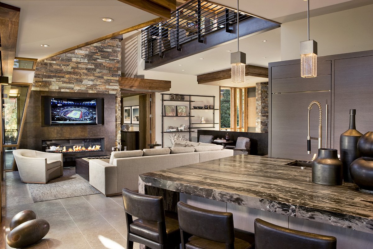 Modern open-concept living room with a stone fireplace, flat-screen TV, beige sectional sofa, and a marble kitchen island with black chairs. Features include wooden accents, shelves with decorative items, and pendant lighting.