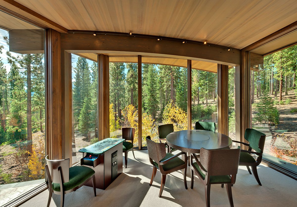 A cozy dining area with six chairs, a round table, and large floor-to-ceiling windows showing a forest view with tall trees and sunlight.