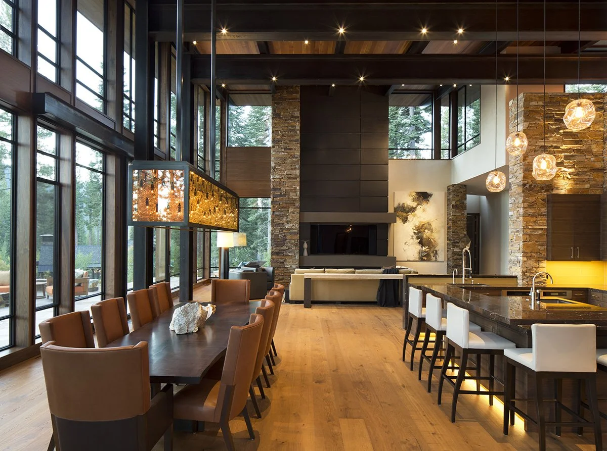 Modern open-concept living and dining area with floor-to-ceiling windows, wooden floors, a large dining table with brown chairs, a kitchen island with white chairs, pendant lights, a stone fireplace, contemporary artwork, and surrounding trees visibl