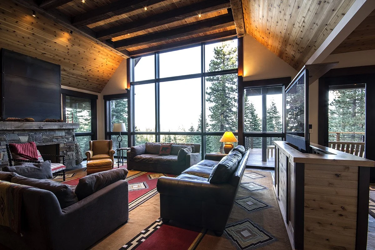 Living room with large floor-to-ceiling windows overlooking a forest, wooden ceiling, stone fireplace, and various sofas and chairs with colorful pillows.