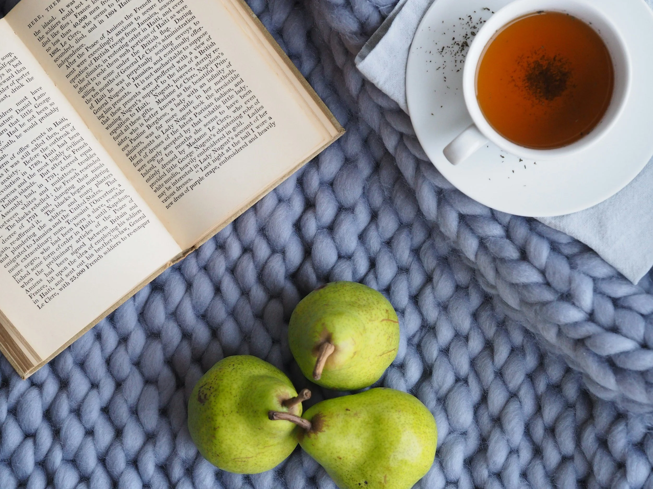 Three green pears, an open book, a white cup of tea on a saucer, and a white napkin on a chunky gray knit blanket.