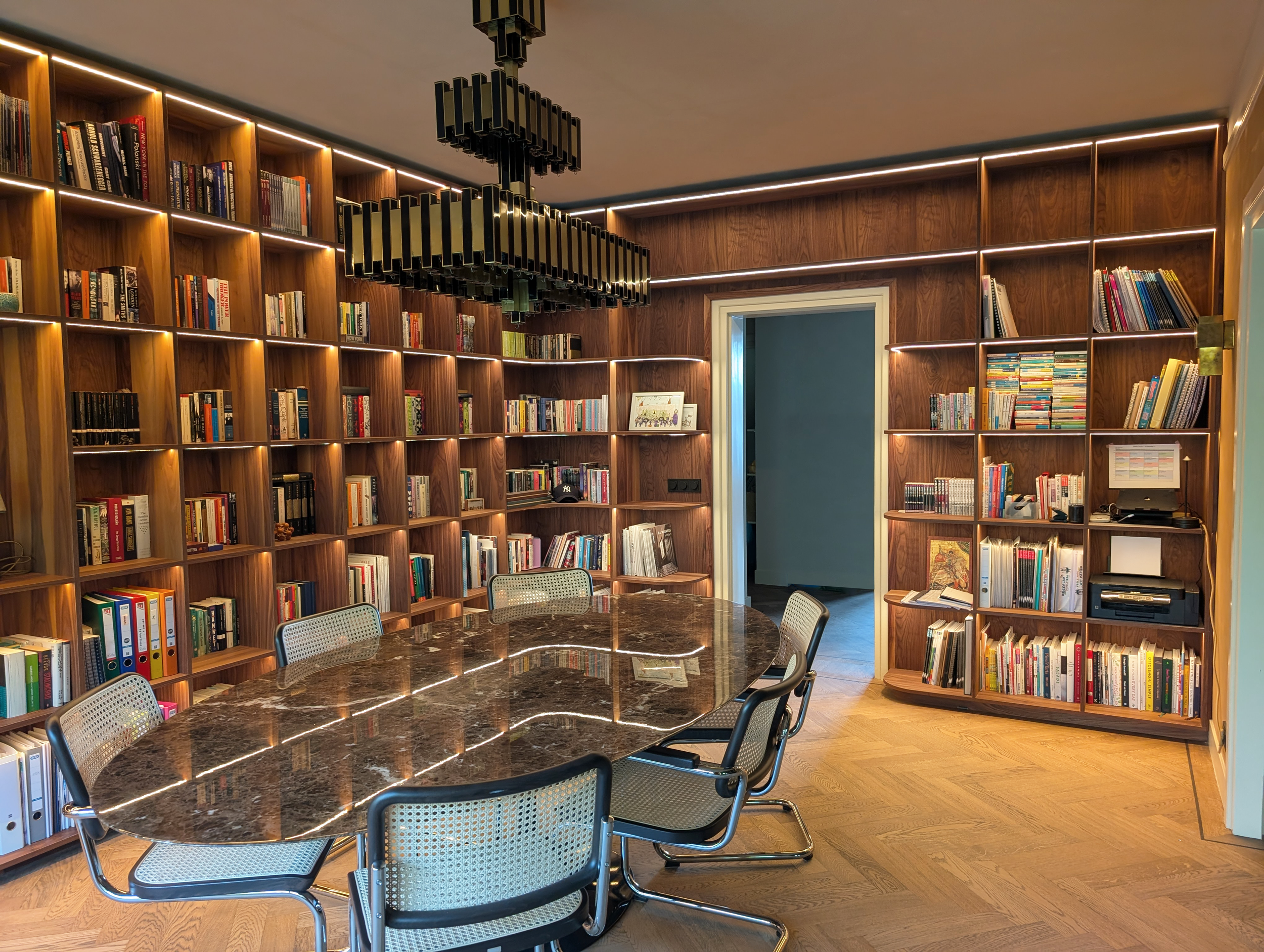 A cozy library room with wooden bookshelves filled with books, a large oval marble-top table, six modern chairs, and a black rectangular chandelier hanging from the ceiling.