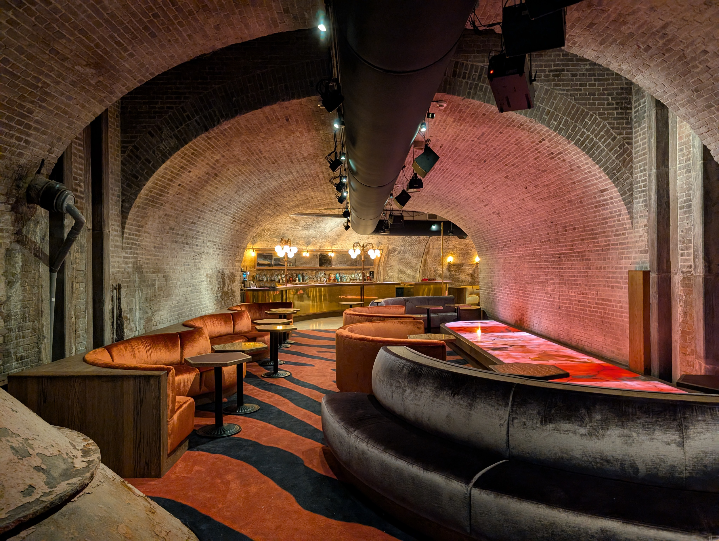 Luxurious underground bar with exposed brick arches, orange velvet seating, and modern bar area with warm lighting.