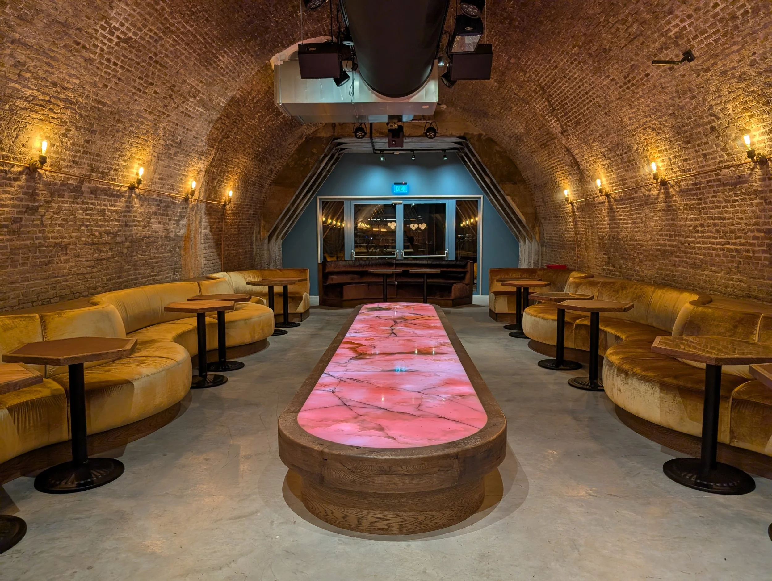 Interior of a stylish lounge with exposed brick arched ceiling, yellow curved velvet couches along the walls, small tables, a central pink marble-topped table, and a stage with blue walls and glass doors at the back.