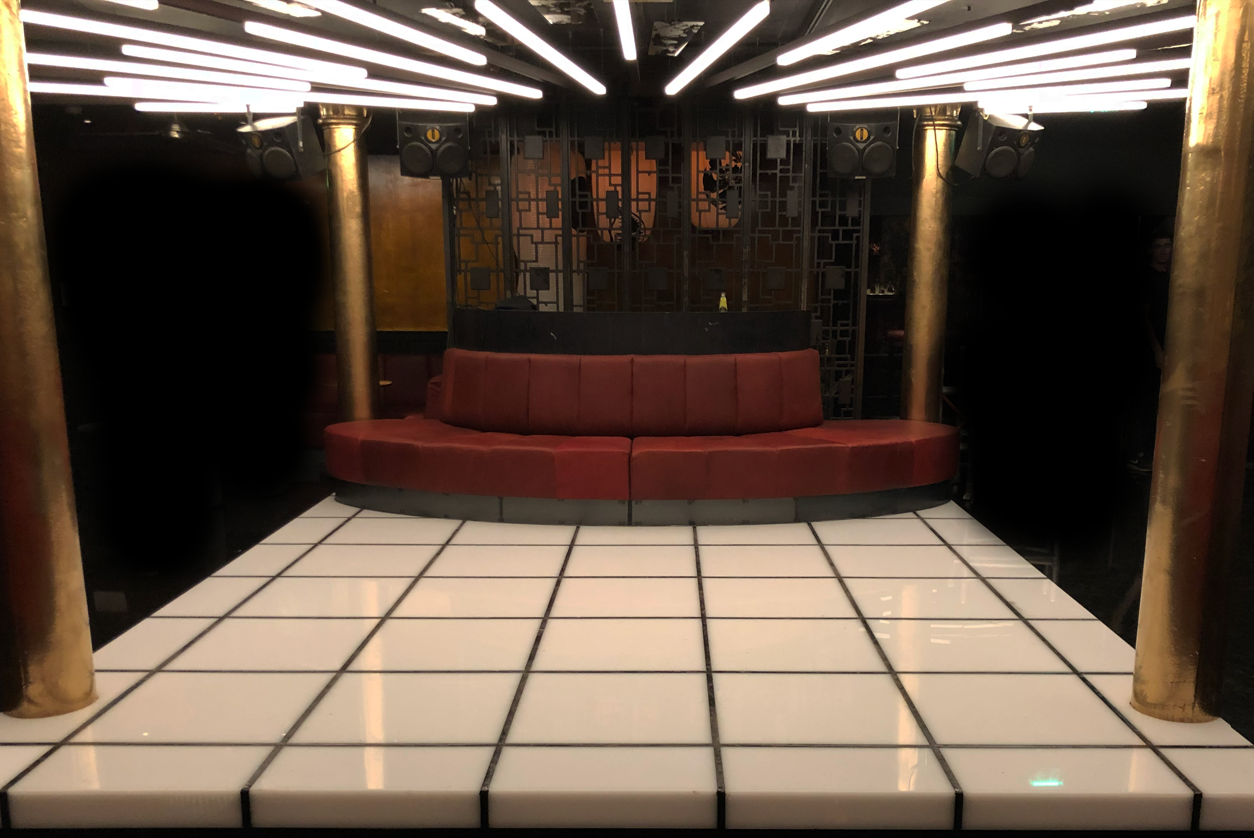 Interior of a nightclub or lounge with a red curved sofa, gold pillars, black decorative wall panel, white tiled floor, and ceiling lights.