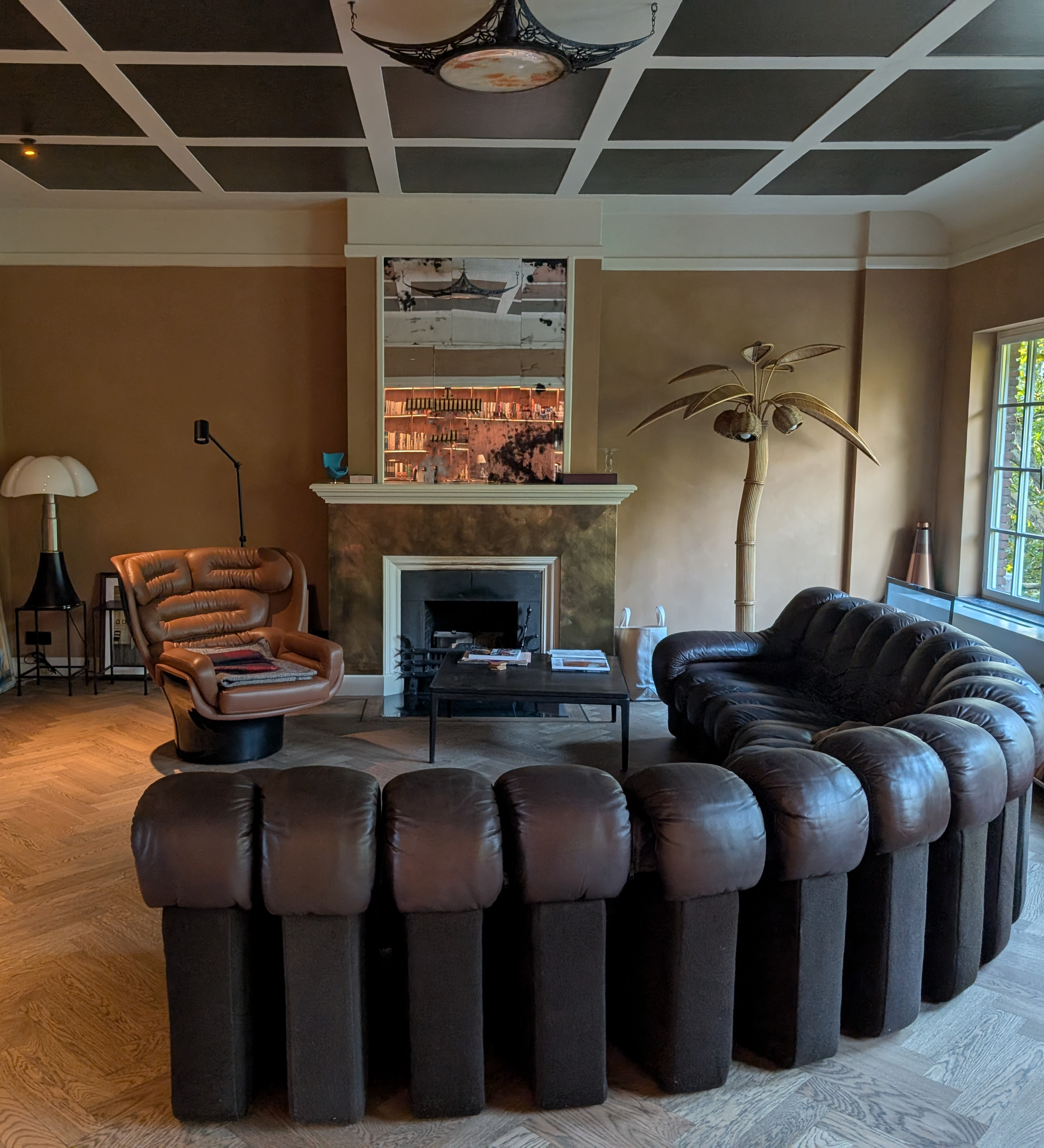 Living room with a brown leather sectional sofa, a tan leather armchair, a black coffee table, a fireplace with a mirror above it, a tall faux palm tree, a window with sunlight, various decorative lamps, and wooden flooring.