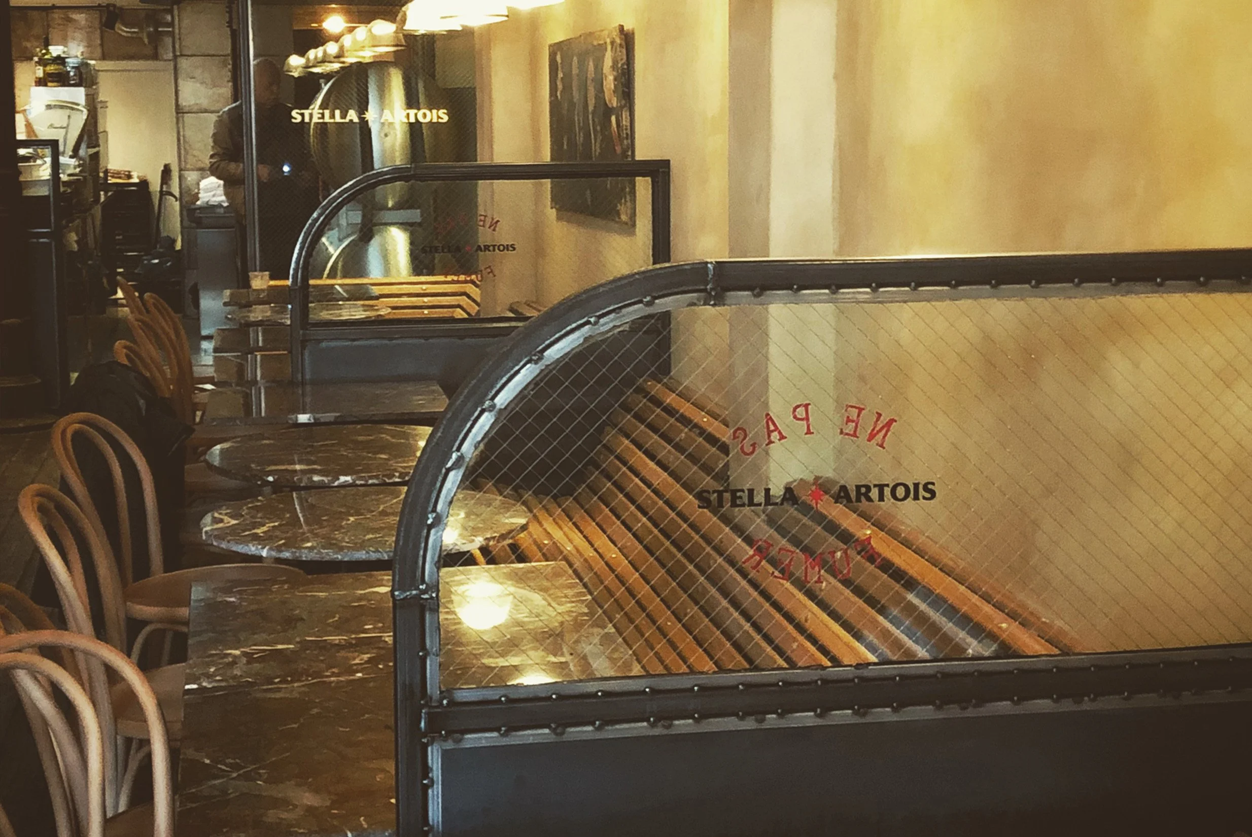 Inside a restaurant with marble tables and wooden chairs, separated by glass partitions with the 'Stella Artois' logo, close to a counter and kitchen area.