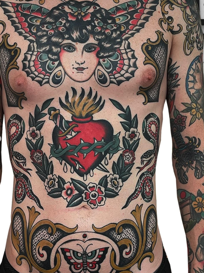 Close-up of a tattooed man's chest with a portrait of a woman with butterfly wings on his upper chest, a sacred heart with a sword, flowers, snakes, and decorative elements.
