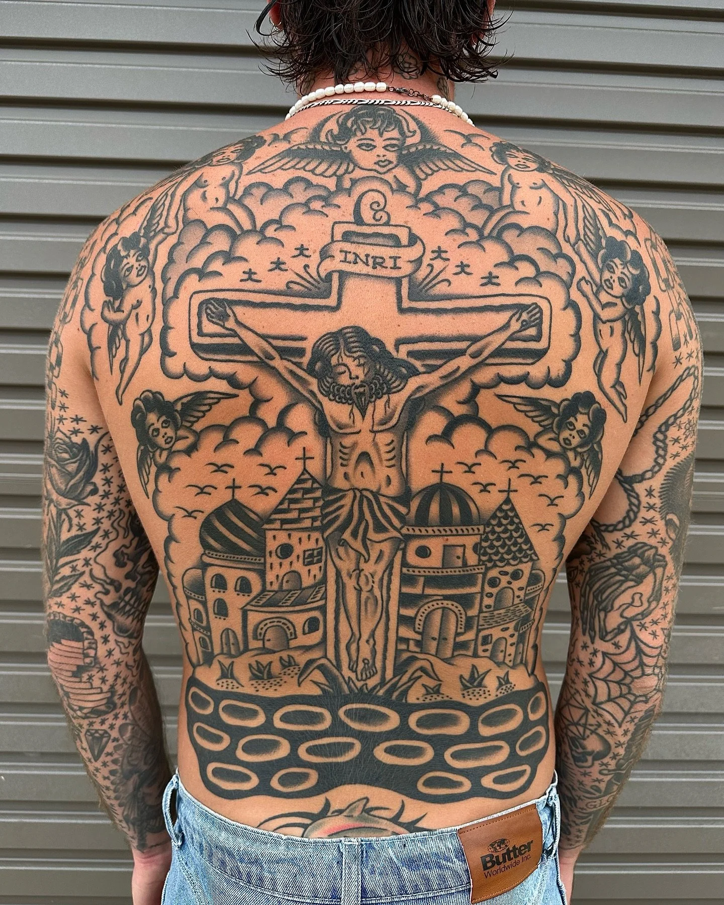 Back tattoo featuring Jesus on the cross, surrounded by clouds, angels, and religious symbols, with a cityscape at the bottom.