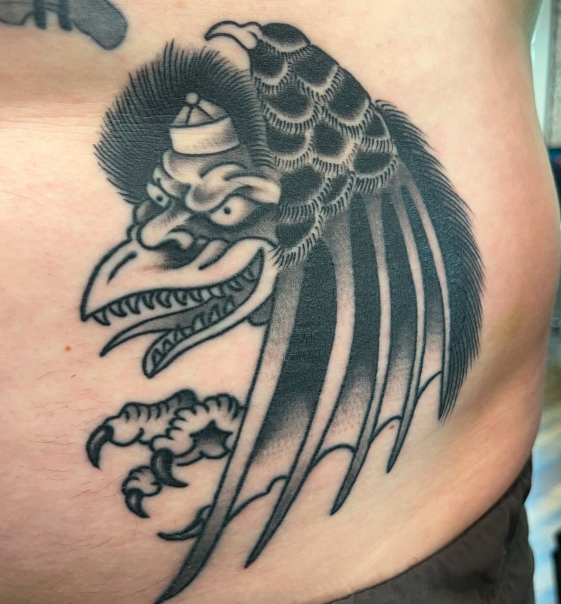 Black and gray tattoo of a mythical creature with the body of a bird and the face of a demon, featuring sharp teeth, claws, and detailed feathers.