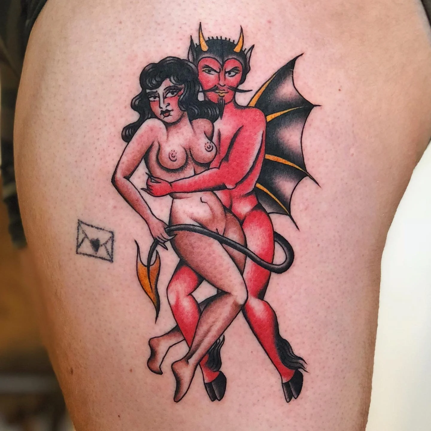 Tattoo of a devil and a woman embracing. The devil has red skin, black wings, and horns, with a tail and a mischievous expression. The woman has dark hair and is nude. There is an envelope symbol nearby.