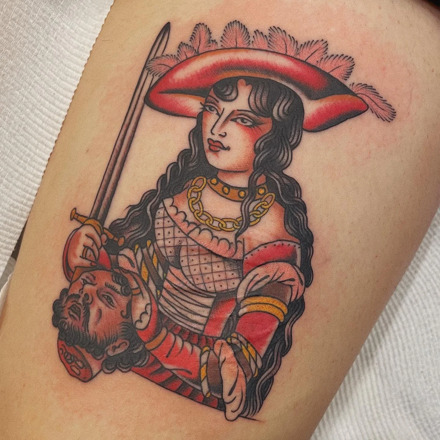 Tattoo of a vintage-style woman with long black hair, wearing a feathered hat, gold chains, and a red and pink outfit, holding a sword and a severed head.