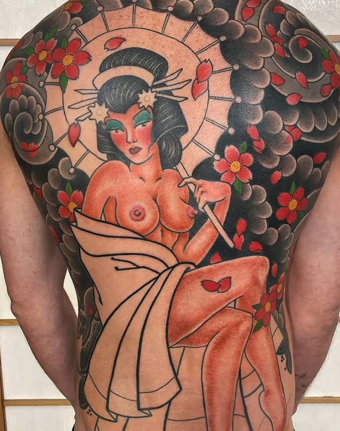 Tattoo of a Malibu Barbie-inspired woman with black hair, green eyes, and pink makeup, surrounded by pink cherry blossoms and black clouds, holding a spear, with a shell headdress and butterfly details.