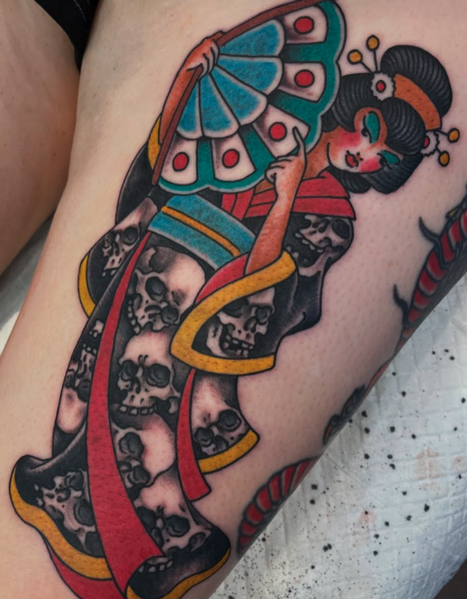Tattoo of a Geisha holding a fan with a butterfly design, surrounded by skulls, with colorful elements including red, yellow, and blue.