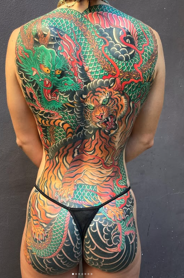 A person with extensive colorful tattoos on their back and lower body, featuring a traditional Japanese-style dragon and tiger battling among clouds and scales.
