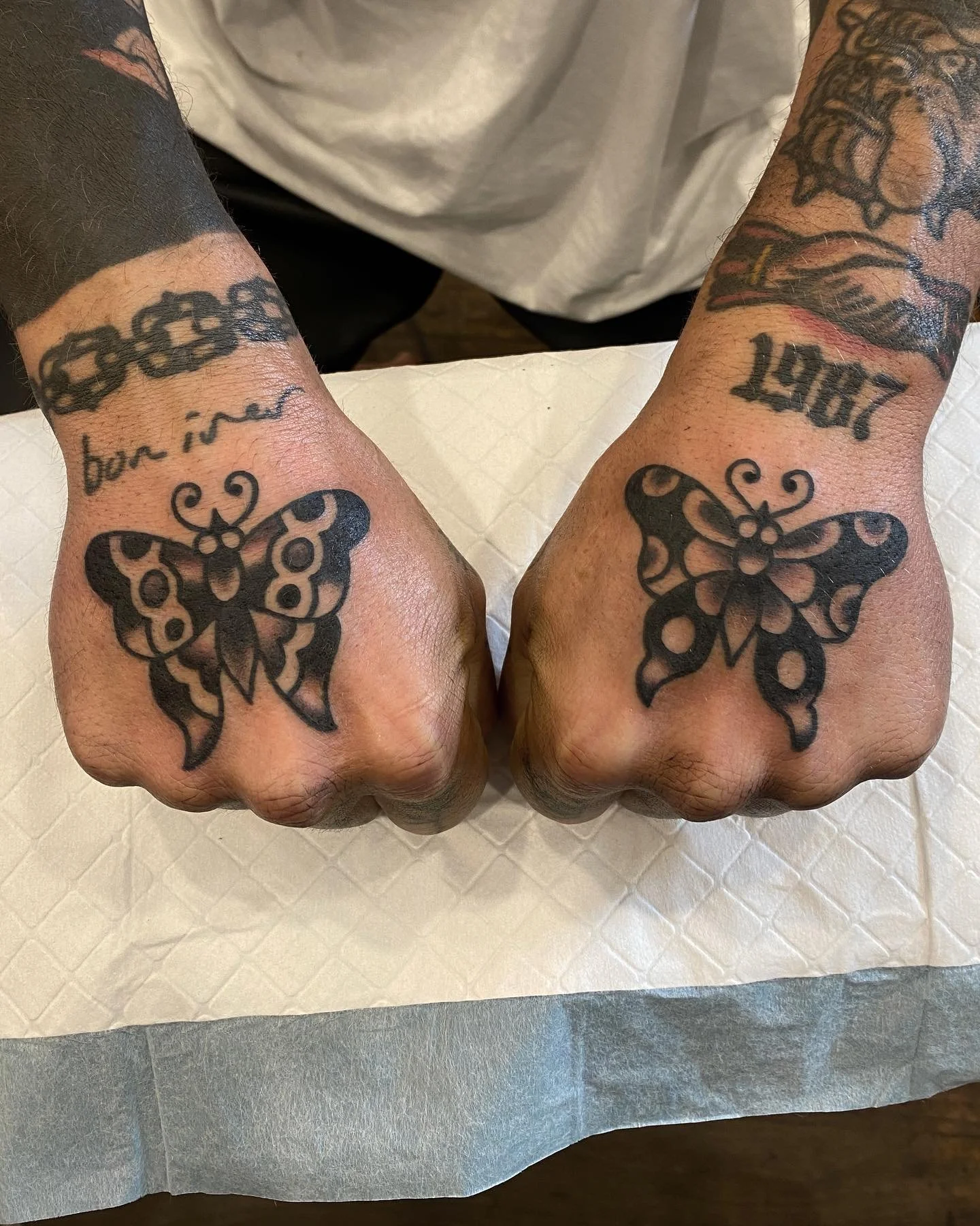 Tattooed fists with butterfly tattoos, the left with a chain tattoo on the wrist and the right with the year 1984 tattooed above the butterfly.