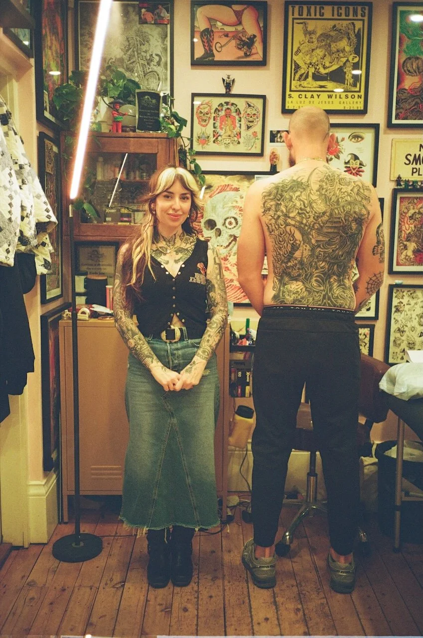 A woman with long hair and tattoos on her arms and neck standing next to a shirtless man with tattoos on his back in an art studio surrounded by framed artwork and tattoo designs on the walls.