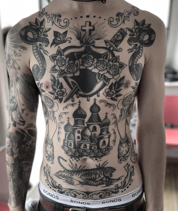 A person's chest and abdomen covered with multiple black and gray tattoos, including snakes, a cross, a dagger, a church, a sleeping tiger, and decorative floral and cloud patterns.