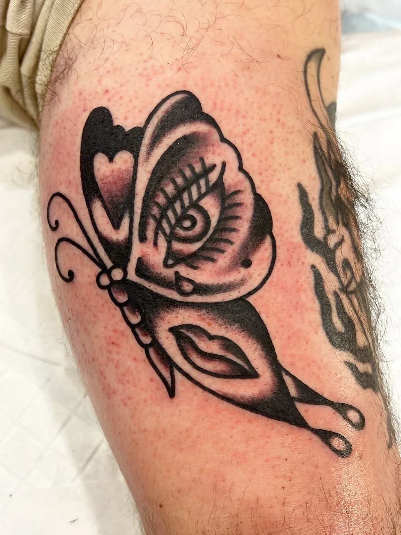 Black outline tattoo of a butterfly with a heart on one wing and a floral design on the other wing, on a person's arm.