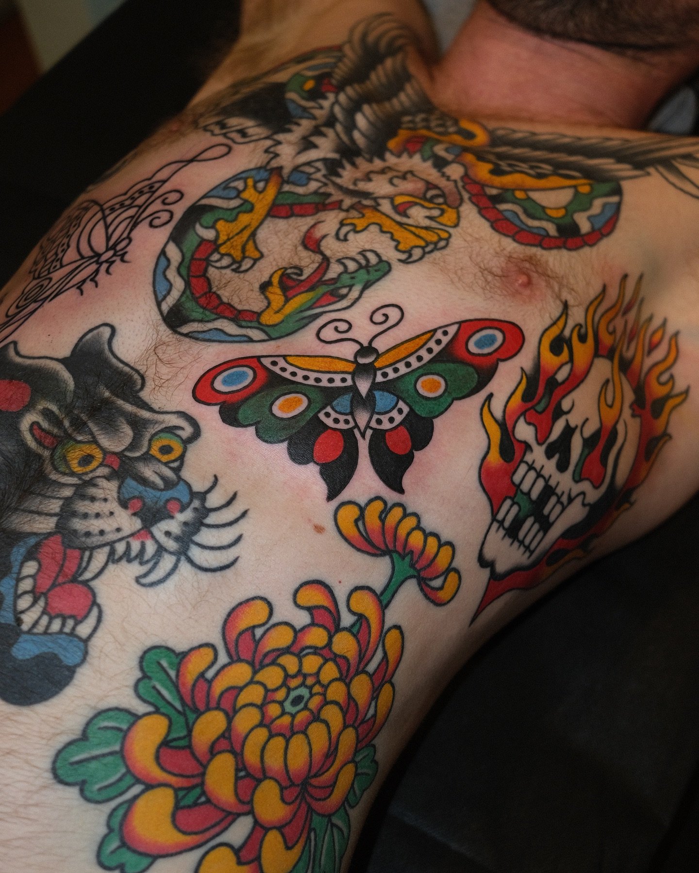 A person's chest with various colorful tattoos, including a butterfly, skull, flower, tiger, and a snake.