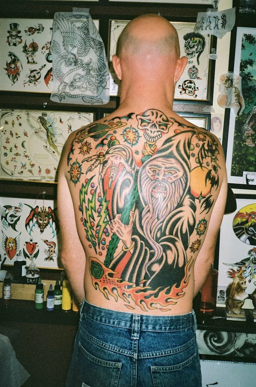 Back tattoo of a wizard with a long beard, holding a staff with a glowing orb, surrounded by stars and mystical elements, with a skull above and colorful lightning bolts.