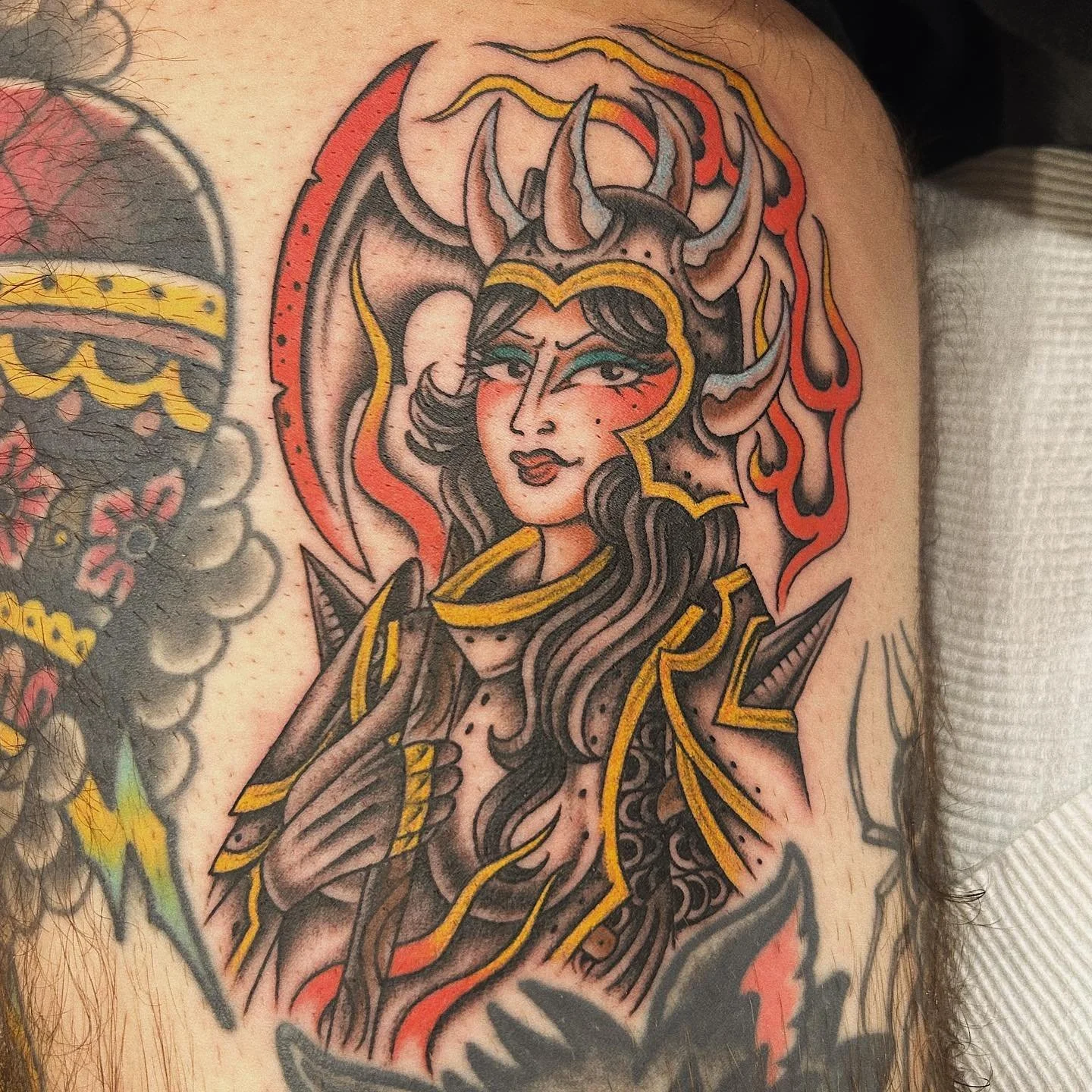Tattoo of a woman with long dark hair, wearing a horned crown and black armor, surrounded by flames, with a confident expression and a wink.