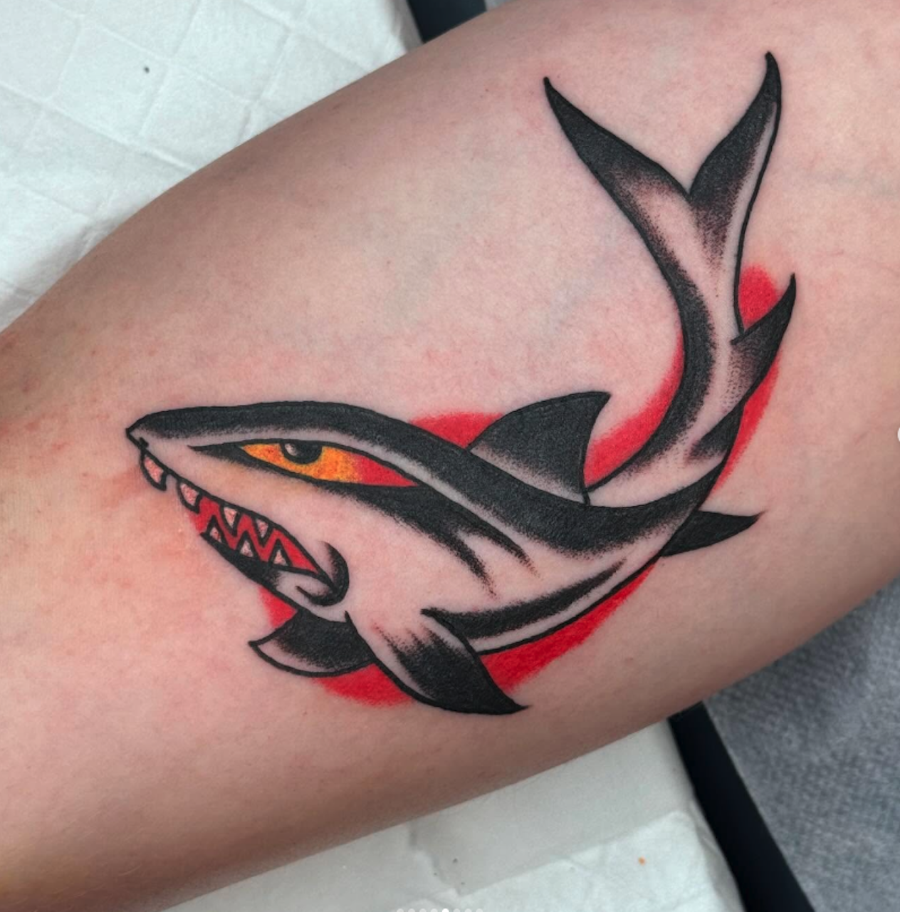 A tattoo of a stylized shark with an aggressive expression, featuring sharp teeth and a dorsal fin, on a person's arm with a red background.