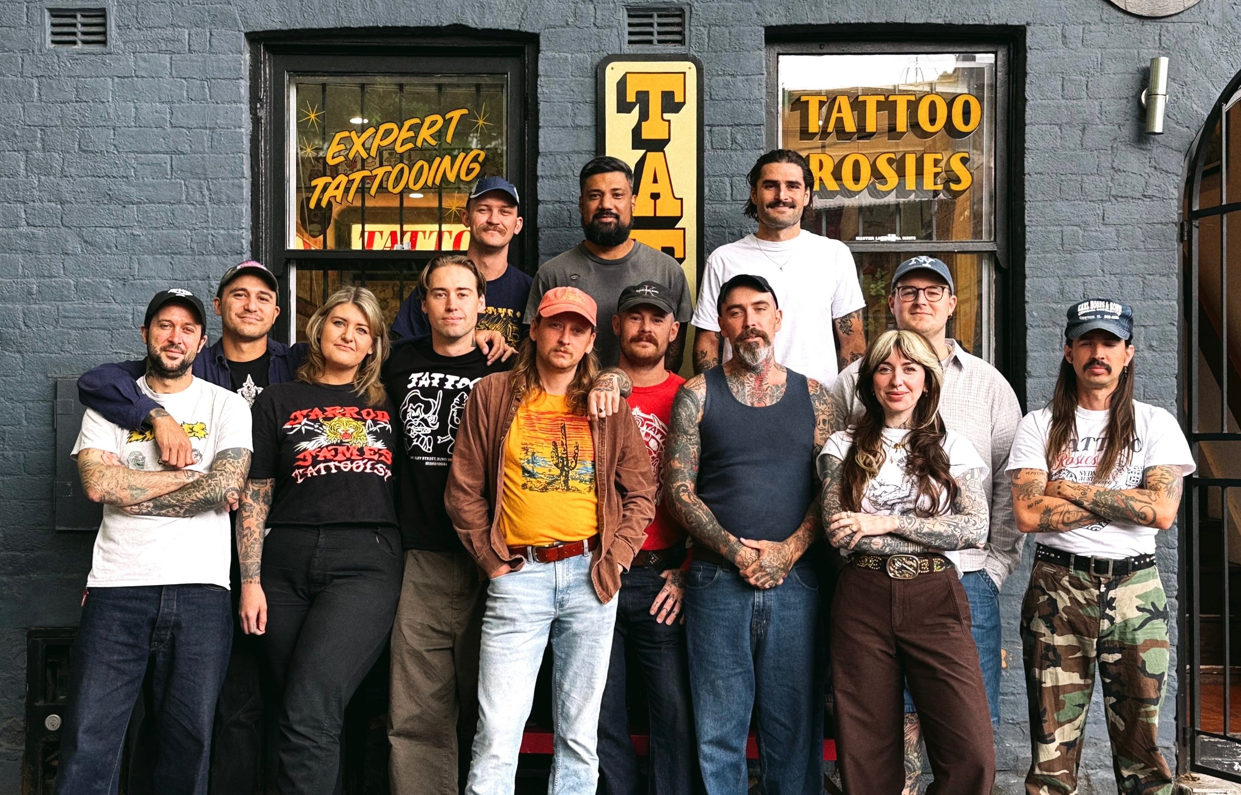Group portrait of tattoo artists standing outside a tattoo parlor, with signs indicating services like expert tattooing and tattoos for rosaries, in front of a dark brick building.
