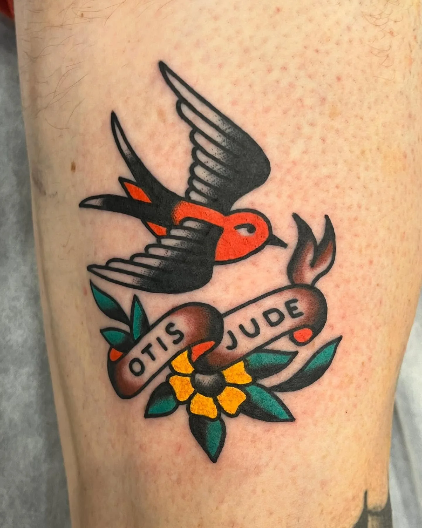 A colorful tattoo of a bird flying over a banner that reads "OIS JUDE," with a yellow flower and green leaves underneath.