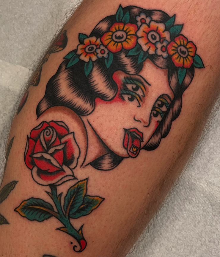 Tattoo of a woman with dark hair wearing a flower crown, with bold lines and bright colors, and a red rose below her face.