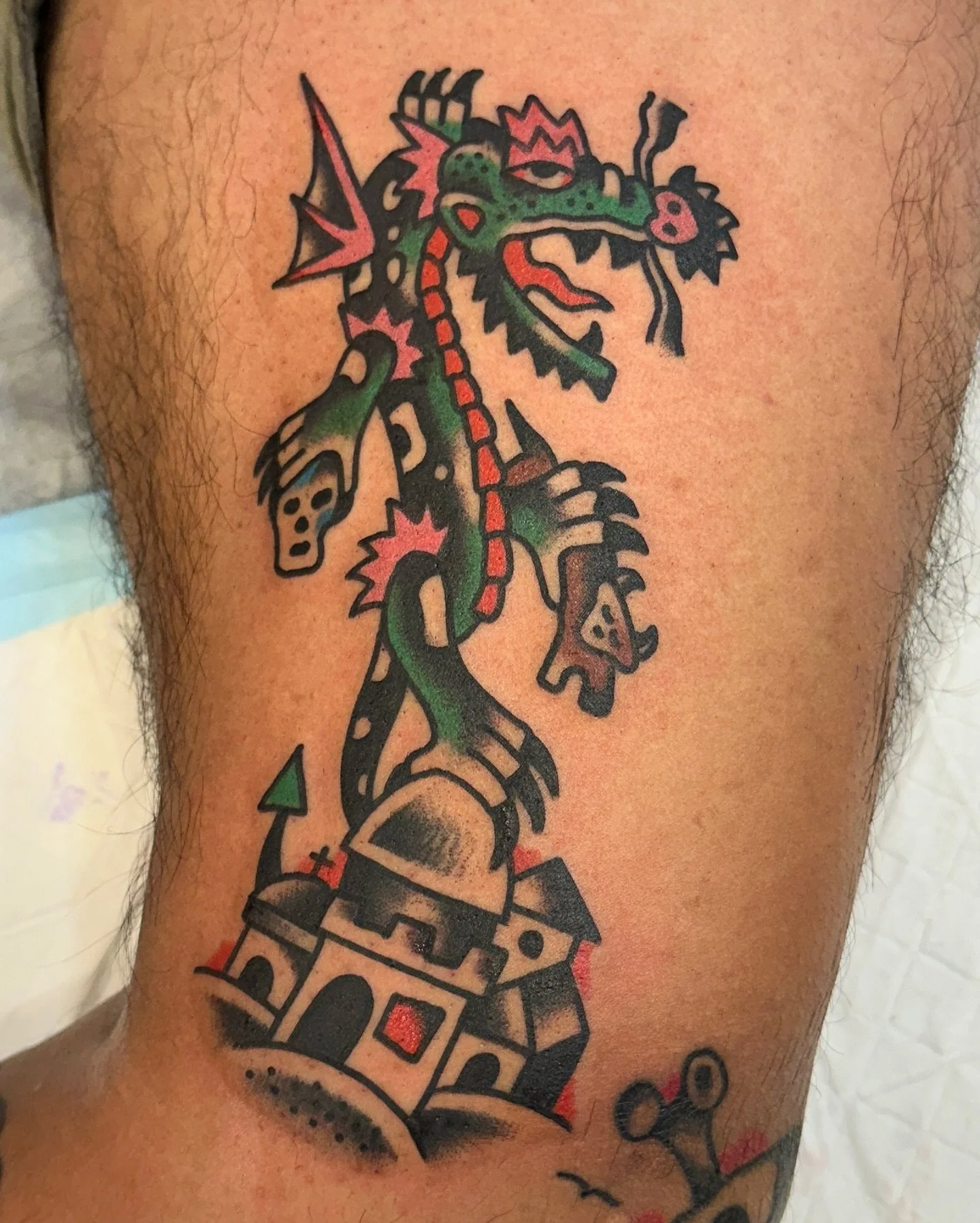 Colorful tattoo of a dragon wrapped around a castle turret, with bold outlines and vibrant green, red, and black colors.