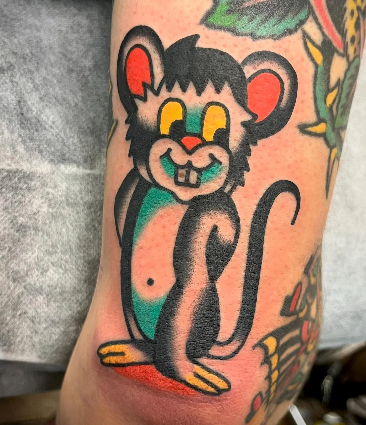 Colorful tattoo of a cartoon mouse, with yellow eyes, a green nose, and wearing a big smile, on a person's arm with other tattoos nearby.