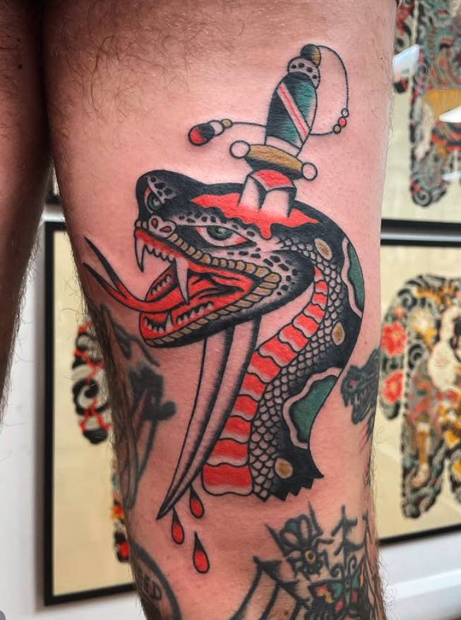Tattoo of a dragon with an open mouth, red tongue, and sharp teeth, with a sword piercing through its head and a dagger and a colorful object above.