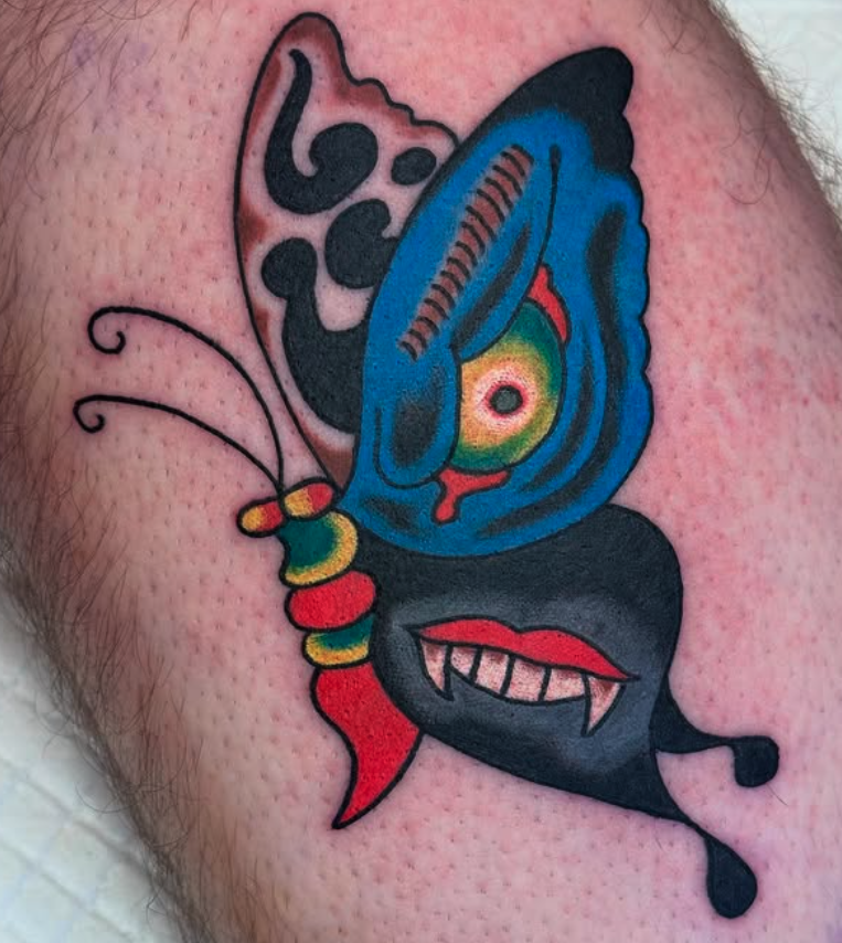 Colorful tattoo of a butterfly with vibrant blue and black wings, red lips, sharp teeth, and detailed antennae on person's skin.