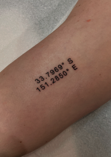 Tattoo of geographic coordinates 33.7696° S, 151.2850° E on a person's arm.